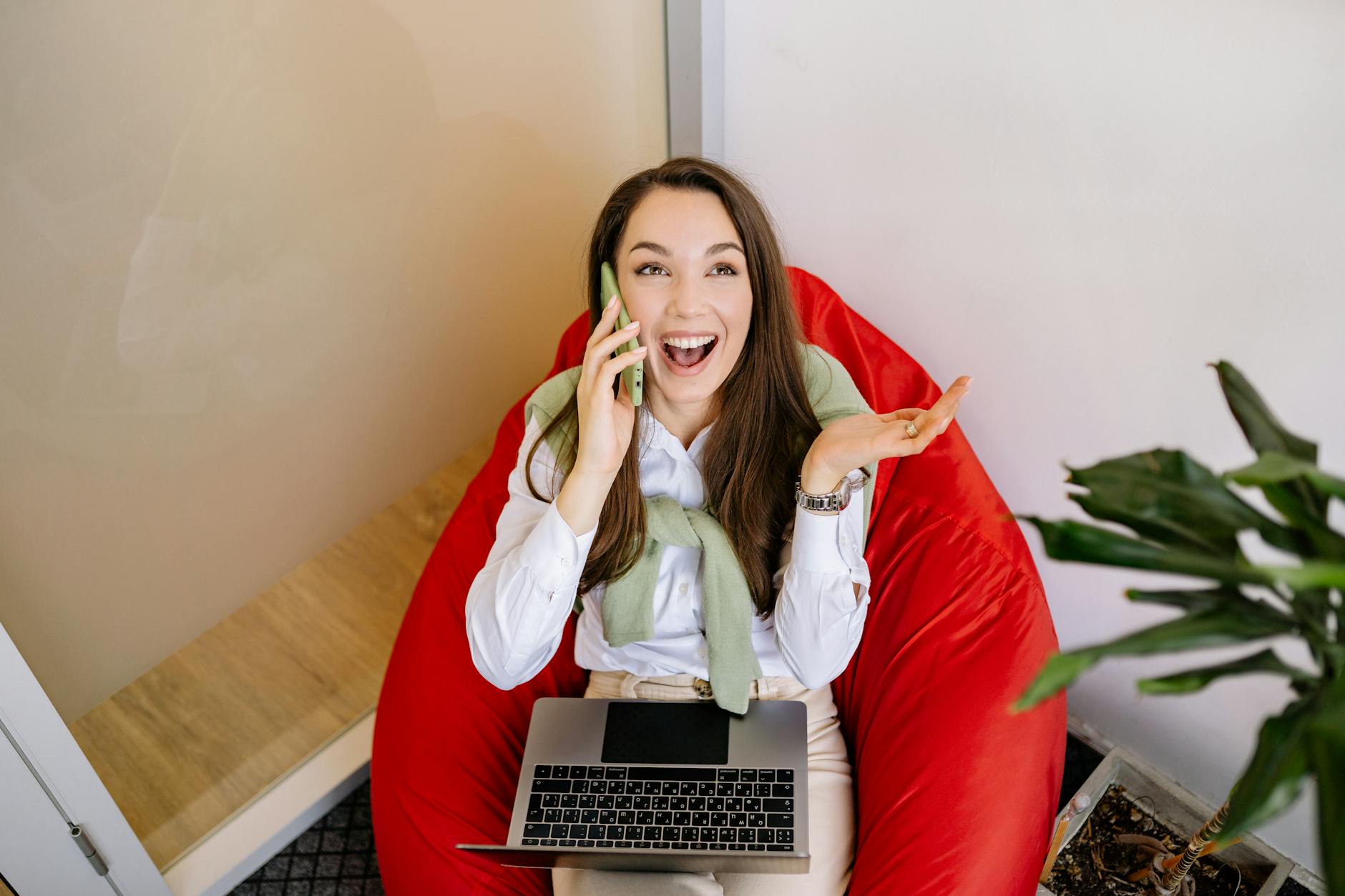 Kobieta rozmawia przez telefon, siedząc na czerwonej pufie sako z laptopem
