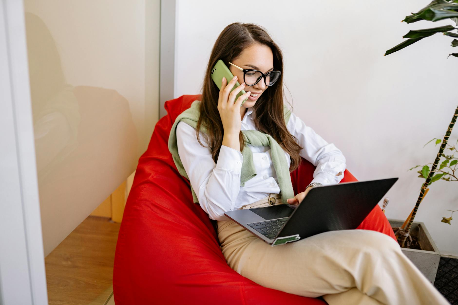 Kobieta pracuje na laptopie i telefonie, siedząc w fotelu w salonie